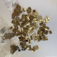 Overview of a large variety of small brass hinges spread out on a white surface with some in plastic bags, showing different shapes and sizes of hinges.