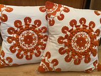 Two down-filled pillows with pale cream covers and tangerine embroidered floral circular designs and tassels, showing front view of both pillows.