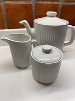 Photo showing the complete tea set comprising of lidded teapot, creamer, and lidded sugar bowl, all in white ironstone with ribbed design.