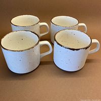 Four ceramic MCM Lifestyle cups with a cream base color and cinnamon speckles shown from the front, highlighting burnt cinnamon rim edges and handles.