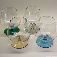 Set of four clear glass brandy snifters with colored feet (green, yellow, blue, clear) side by side on flat surface
