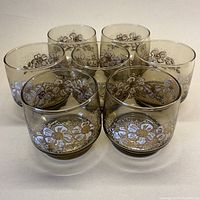 Seven smoky grey glasses with white and gold floral enamel decoration arranged in a group, showing front and side views of the floral design.