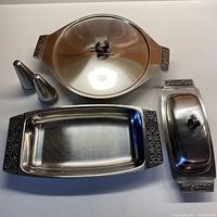 Top-down view showing the entire set of mid-century stainless steel servingware with decorative raised and blackened floral scroll details on handles and lids.