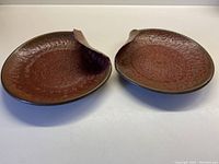 Two pottery serving plates showing clay and cinnamon hues with milk chocolate border, slightly bumpy textured surface, raised circular detail and scalloped bent edge.
