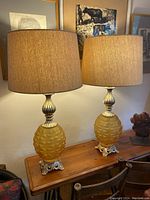 Two ornate mid-century lamps with amber textured glass bodies and cream fabric shades standing on a wooden table against a wall with framed artwork.