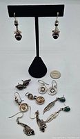 Collection of various pairs of silver earrings on a display stand with quarter for scale. Includes earrings with black stones, embossed round details, and feather designs with green stones.
