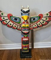 Front view of the hand carved totem pole showing the bird figure with bright painted wings and multiple carved faces on the pole.