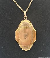 Close-up of front of gold filled Art Deco locket on chain against black background, showing geometric ribbed design with scalloped edges and central initial monogram