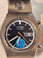 Front view of watch dial showing black face, orange chronograph hand, blue subdial, day-date window.