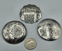 Photo showing all three round vintage sterling silver brooches with floral scrollwork engravings, next to a coin for scale.