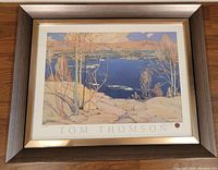 Framed print showing winter landscape scene with bare trees, water, and hills, signed with Tom Thomson name and wax seal below the artwork.