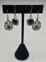 Front view of both pairs of earrings on black display stand showing red oval stone pair with silver filigree, and dark blue round stone pair with leaf design.