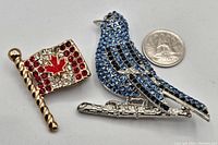 Top view of both rhinestone brooches next to a quarter for scale: bluebird with blue and black rhinestones and Canadian flag with red and clear rhinestones and red maple leaf.
