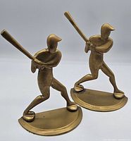 Front view of two brass baseball player bookends showing batting pose, detail of helmet, bat, and base.