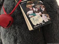 Close-up of gray teddy bear showing quality mohair fabric, red jingle bell necklace, and original price tag of $40, with booklet mentioning 'One More Bear' by Cindy Anschutz.