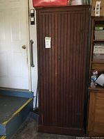 Full view of tall wooden cabinet with vertical slat door closed, placed beside steps and next to a desk in a garage.