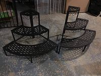 Two black metal corner display shelves with three tiered curved lattice shelves, arranged side by side on floor.