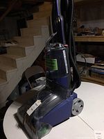 Full view of the upright carpet cleaner on a round white table in basement setting, showing blue and black plastic body with rear wheels and upright handle.