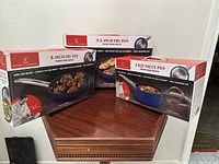 Three boxes of Emeril Lagasse cookware: 8-inch fry pan, 9.5-inch fry pan, and 1.5 QT sauce pan, arranged on a wooden surface.