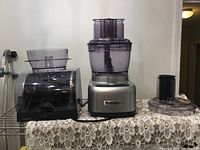 Photo showing Cuisinart food processor motor base, two processing bowls with lids and feed tubes, and black plastic base for attachments on lace tablecloth.