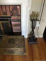 Fireplace tool set stand with poker, shovel, tongs with brass finials next to decorative metal overlay wood storage box on floor by fireplace.