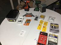 All items spread on white table showing two cordless drills, sanding blocks, hardware boxes, and other supplies.
