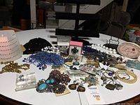 Overview of assorted jewelry pieces including colorful necklaces, bracelets, earrings, crystal and bead strands, jewelry boxes, and a black sequin beanie hat, all spread on a white table.