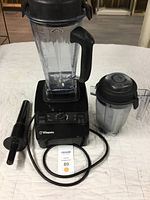 Vitamix motor base on white surface with large blending container, dry container, tamper, and power cord visible