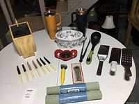 Full view of kitchen utensils and items on a white table including knives, spatulas, ladle, graters, apple slicer tool, porcelain Dutch oven, thermal mugs and drawer liners.