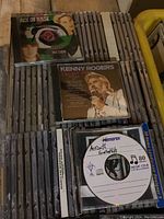 Photo of multiple stacks of music CDs including Ace of Base The Sign and Kenny Rogers Greatest Hits visible on top. CDs are in jewel cases and placed in a yellow plastic bin.