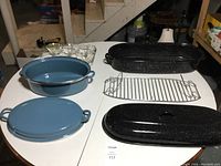 The full set displayed on a table in a basement, showing blue enamel Dutch oven with lid, black speckled roasting pan with lid, wired rack, and glass custard cups in rack.