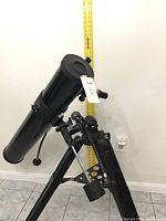 Large black reflector telescope and mount positioned against wall with measuring tape indicating height.