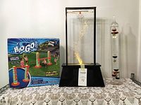 Boxed H2O Go limbo sprinkler, bubble lava lamp, and Galileo thermometer on lace tablecloth