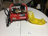 Full view of the red Craftsman electric air compressor with yellow coiled air hose lying on a white table.