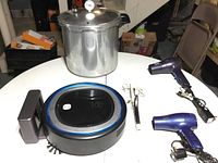 All items in the lot on a white round table, visible pressure cooker, two hair dryers, curling iron, robot vacuum and docking station.
