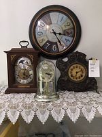Four clocks displayed together on a lace cloth. Includes large wall clock with multicolor numerals, Baroque style dark ornate clock with exposed hands, wooden mantel clock with brass elements (Seth Thomas style), and anniversary clock with glass dome.