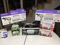 Photo showing various packages of LED light bulbs from Enhance, MaxLite, and Feit Electric brands, arranged on a table in basement environment. Packages include 6-pack and 2-pack boxes, mainly standard LED bulbs and spotlight varieties.