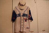 Front view of Blue Jays jersey with cap hung on a wall, showing full details of text, patches, and buttons.