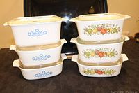 Stacked view of three Blue Cornflower and three Spice o' Life Corningware dishes with glass lids showing decal designs on the sides