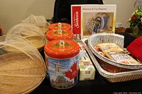 Photo shows assorted kitchen items including bamboo baskets with mesh covers, cookie containers, orange lidded tins with Canadian imagery, and a boxed Sunbeam bakeware and tray organizer.