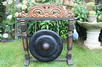 Front view of the large black metal domed gong suspended in carved wooden frame in garden setting.