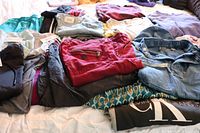 Mixed clothing lot laid out on bed including jackets, pants, and tops in multiple colors and fabrics