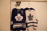 Blue Toronto Maple Leafs captain's jersey with white practice jersey and grey toque hanging on a rod.