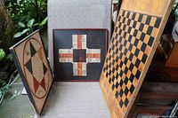 Three vintage wooden game boards - Chinese checkers, Parcheesi, and checkers - displayed on a chair, showing wear and painted details