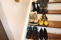 Full lot view showing 6 pairs of women's boots on stairs comprising different styles and colors including black, beige, yellow, brown and dark blue boots.