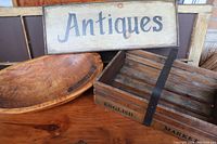Photo of the carved wooden bowl, English market basket, and rectangular antiques sign placed on wooden table.