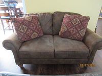 Front view of brown Broyhill love seat with two large patterned pillows on seat cushions, set on wooden floor.