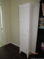 Front view of tall white wooden storage cabinet with two slatted doors closed, showing narrow and tall design, placed against a wall.