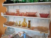 Wide view of shelves with various pink and amber carnival glass items including bowls, plates, dessert cups, and bottles.