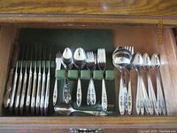 Set of matching stainless steel flatware including knives, forks, tablespoons, serving spoon and ladle arranged neatly in a drawer with green felt lining